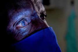 Mann mit Mund-Nasen-Schutz im Dunkeln, eine Lampe beleuchtet seine rechte, weiß schimmernde Pupille