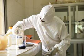 Ein weiß gekleideter Mann mit Mundschutz gießt in einem Labor eine Flüssigkeit in ein Glasgefäß.