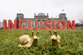 Zwei goldene Schnecken sitzen im Gras vor dem Bundestag in Berlin. Im Hintergrund steht das Wort &quot;Inklusion&quot;.