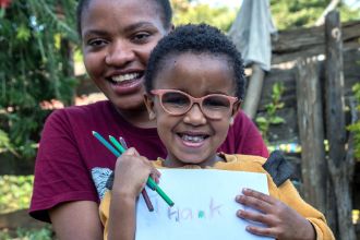 Eine Frau und ein lachendes Kind mit Brille. Es hält ein Blatt mit der Aufschrift &quot;Thank u&quot; in der Hand.