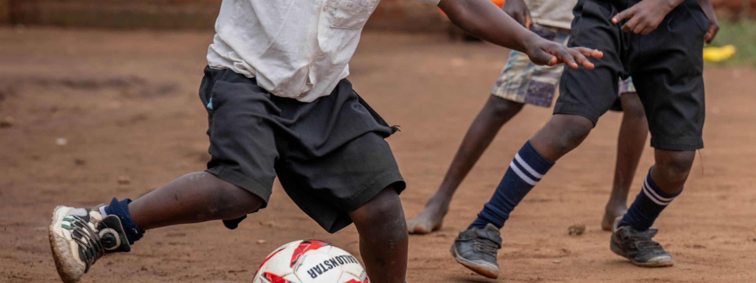 Drei etwa siebenjährige Jungen spielen Fußball in einem von einer Backsteinmauer begrenzten Hof.