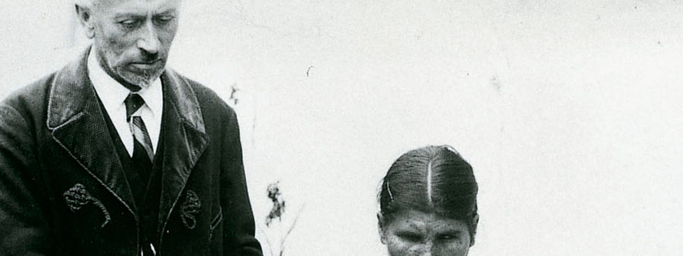 Ein Mann im Anzug, wie man ihn um 1925 trug, steht neben einem Tisch, an dem drei junge Frauen sitzen. Diese tasten mit den Händen über die Seiten großer, aufgeschlagener Bücher.