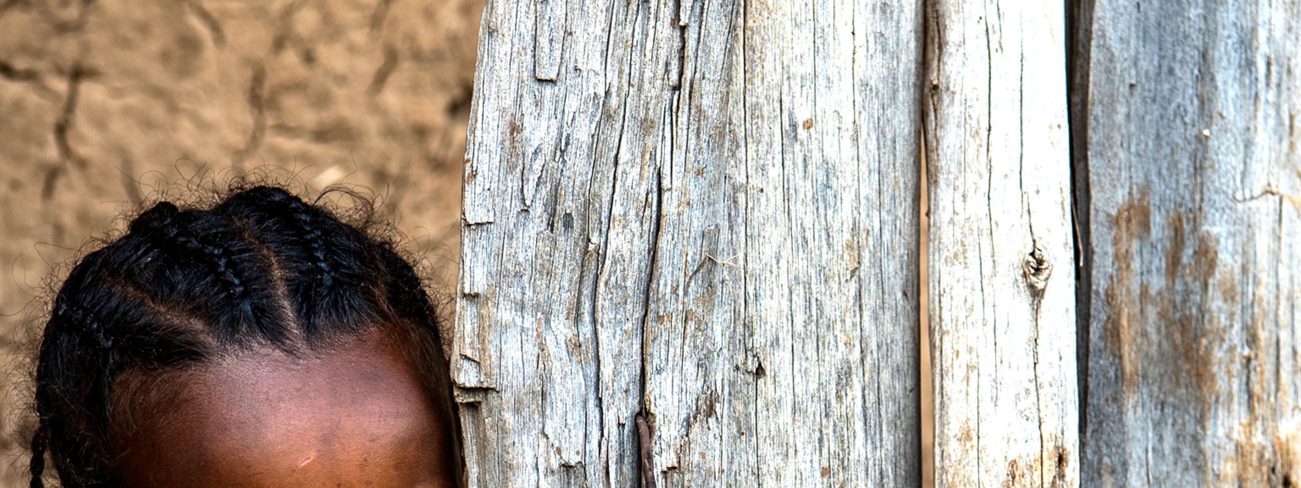 Ein etwa fünfjähriges Mädchen mit eng am Kopf anliegenden Zöpfchen schaut hinter einer Bretterwand hervor.