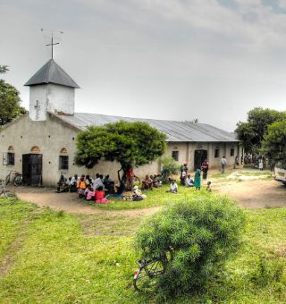 Landschaft mit Kirche, vor der eine Gruppe Menschen in bunter Kleidung sitzt.