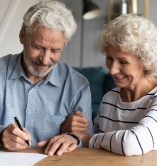 Testamentsspende: Ein älteres Paar sitzt am Tisch. Der Mann hält einen Stift und schreibt auf ein Blatt Papier seine Nachlassspende.