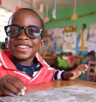 Ein etwa dreijähriger Junge mit einer großen Brille sitzt an einem Tisch im Klassenzimmer. Er lacht.