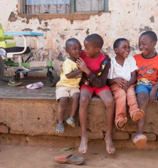 Vier afrikanische Kinder sitzen vor einer Hütte, im Hintergrund ein Rollstuhl