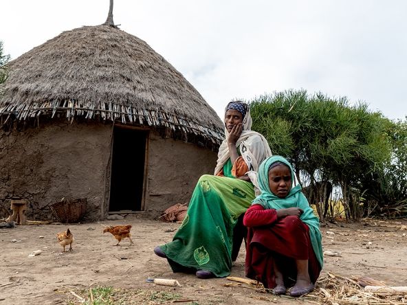 Etwa etwa neunjähriges Mädchen und eine Frau sitzen vor einer Rundhütte. Hühne laufen um sie herum.