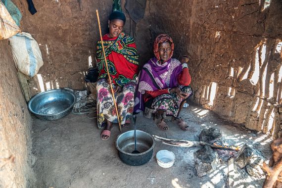 Eine junge und eine ältere Frau sitzen in einer Lehmhütte, vor ihnen steht ein leerer Kochtopf.