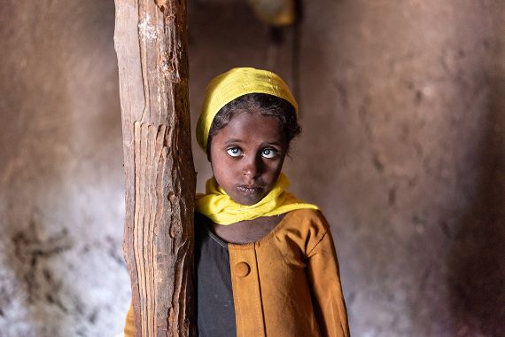 Ein etwa fünfjähriges Mädchen mit gelbem Kopftuch und weißen Pupillen steht an einem Holzpfosten vor einer Lehmwand.