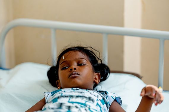 Ein etwa fünfjähriges Mädchen liegt in einem Krankenhausbett. Über dem rechten Auge hat es ein kleines, weißes Markierungspflaster.