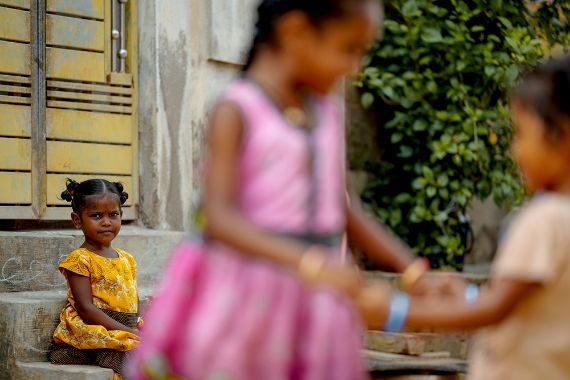Ein etwa fünfjähriges Mädchen mit kleinen dunklen Zöpfchen sitzt auf einer Treppe. In seiner Nähe spielen Kinder.