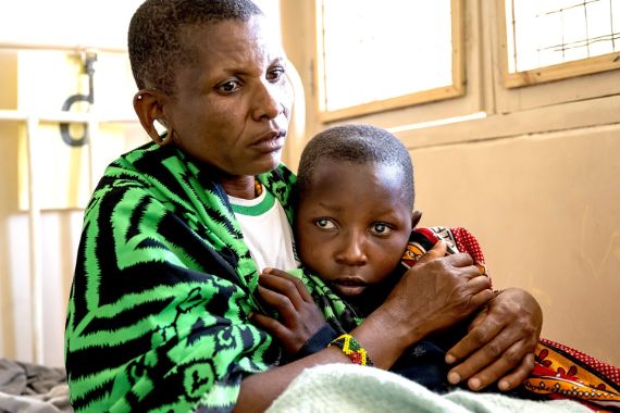 Ein etwa zehnjähriges Mädchen mit hellen Pupillen schmiegt sich an eine Frau in Massai-Tracht.