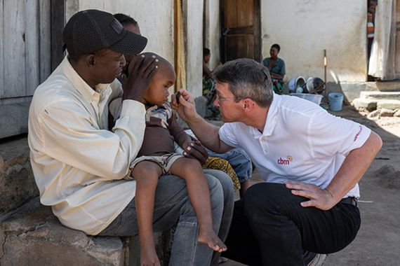 Ein Mann untersucht mit einer kleinen Taschenlampe die Augen eines etwa zweijährigen Jungen, der auf dem Schoß eines anderen Mannes sitzt. 