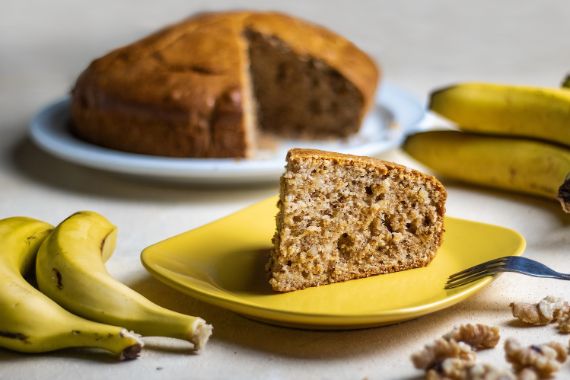 Ein Telle mit einem Stück Kuchen, auf dem zwei Bananenscheibchen liegen. Neben dem Kuchen liegt eine Kuchengabel.