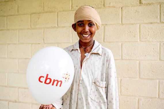 Ein Junge mit Kopfverband hält einen Luftballon in der Hand, auf dem &quot;CBM&quot; steht.