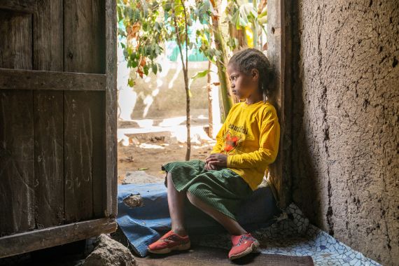 Ein etwa neunjähriges Mädchen sitzt auf der Türschwelle einer Hütte.