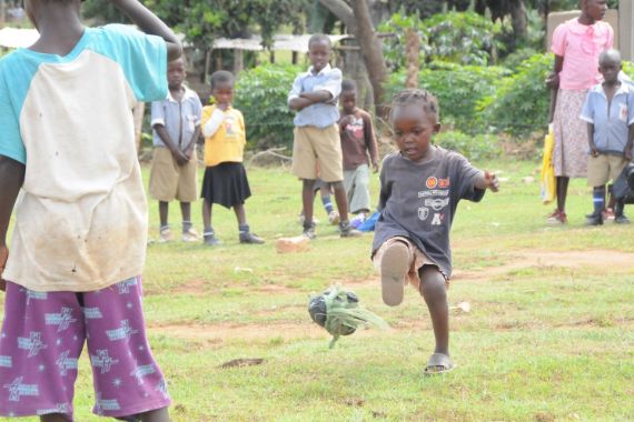 Ein kleines Mädchen kickt einen Ball, andere Kinder und Erwachsene schauen zu.