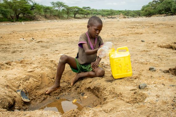 Ein etwa siebenjähriger Junge sitzt vor einem Wasserloch in einem ausgetrockneten Flussbett und schöpft Wasser in einen Plastikkanister.