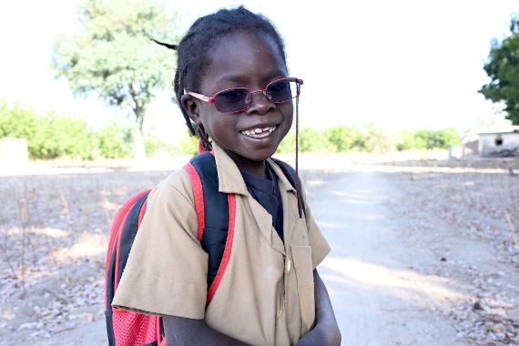 Ein lachendes, etwa achtjähriges Mädchen mit Brille trägt einen Rucksack und eine beige Schuluniformbluse.
