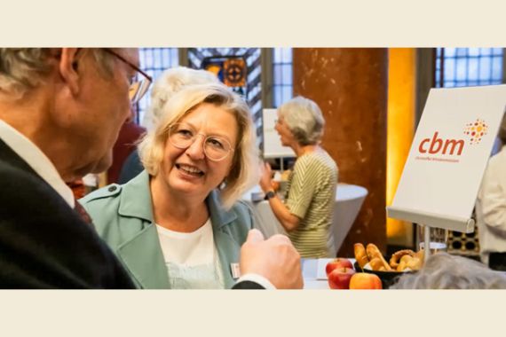 Eine Frau im Gespräch mit einer weiteren Person vor einem Tisch mit Äpfeln und Gebäck und einem Poster mit CBM-Logo.