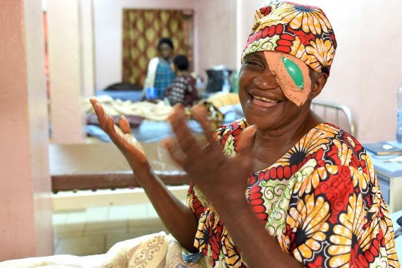 Eine buntgekleidete afrikanische Frau mit Augenverband hält lachend die Hände hoch.