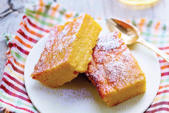 Zwei viereckige, gelbe, mit Puderzucker bestreute Kuchenstücke auf einem weißen Porzellanteller. Dieser steht auf einer buntkarierten Serviette.