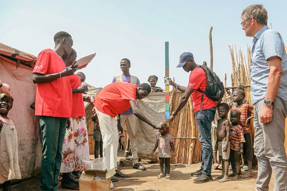 Zwischen zwei Hütten stehen Erwachsene und Kinder um ein etwa dreijähriges Kind. Ein Mann hält eine buntbemalte Messlatte hinter das Kind, ein anderer beugt sich zum Kind hinunter und gibt ihm etwas zu trinken.