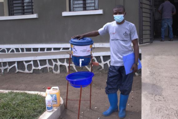 Ein Mann steht neben einem kleinen Wassertank mit Zapfhahn. Darunter eine Schüssel, direkt daneben eine Flasche mit Desinfektionsmittel.