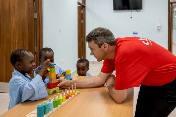 Ein Mann beugt sich über einen Tisch auf dem Spielzeug liegt und spielt mit Kindern, die ihm gegenübersitzen.