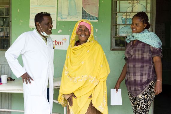 Mann im weißen Kittel, lachende Frau im gelben Gewand und eine weitere Frau, die Papiere in der Hand hält.