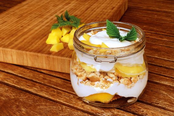 Glas mit Deckel, gefüllt mit weißer Creme, Mangostückchen und Keksbröseln in Schichten.