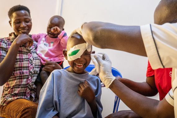 Ein Mann entfernt die Pflaster von den Augen eines sechsjährigen Kindes. Neben dem Kind sitzt eine Frau mit einem ca. einjährigen Kind auf dem Arm. 