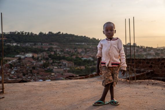 Ein Junge mit X-Beinen steht auf einem Dach. Im Hintergrund sieht man die Stadt Kampala.