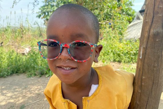 Donor online: donate for children like Ahmed! – A small boy with sunglasses leans against a wooden post.