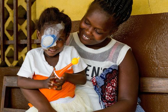 Kleines afrikanisches Mädchen mit Augenverband auf dem Schoß einer Frau