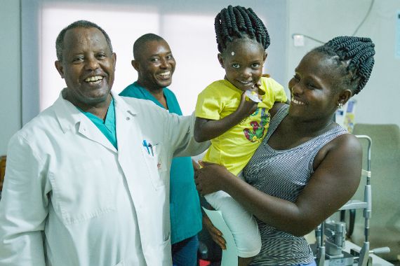 Kleines afrikanisches Mädchen auf dem Arm einer Frau, daneben ein Mann in weißem Kittel