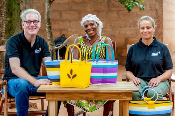 Ein Mann und zwei Frauen sitzen an einem Holztisch, auf dem bunte Taschen mit langen Henkeln stehen. (Spendenprojekt zur Förderung von Frauen mit Behinderungen in Togo)