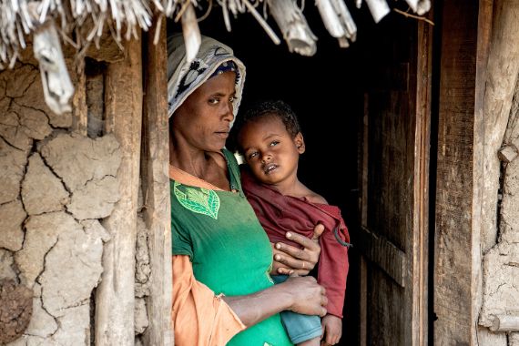 Eine etwa 35-jährige Frau mit einem etwa dreijährigen Kind auf dem Arm steht im Eingang einer strohgedeckten Lehmhütte.