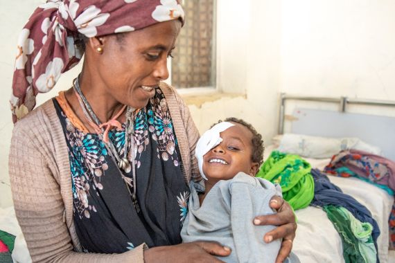 Eine etwa 35-jährige Frau mit einem etwa dreijährigen Kind auf dem Arm steht im Eingang einer strohgedeckten Lehmhütte.