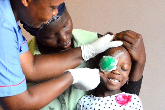 Eine afrikanische Frau im blauen Kittel entfernt den Augenverband eines kleinen Mädchens.