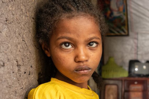 Eine etwa 9-jähriges Mädchen lehnt sich an die braune Lehmwand eines kleinen Zimmers, in dem ein Sideboard und ein niedriger Tisch steht.
