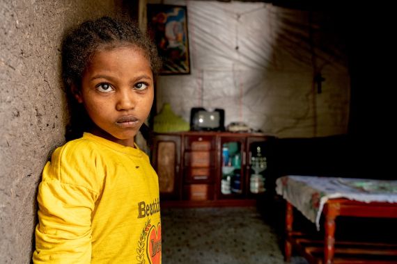 Eine etwa 9-jähriges Mädchen lehnt sich an die braune Lehmwand eines kleinen Zimmers, in dem ein Sideboard und ein niedriger Tisch steht.