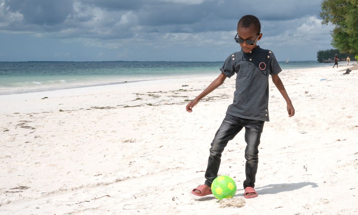 Ein Junge mit dunkler Brille läuft am Strand und kickt einen grünen Plastikfußball.