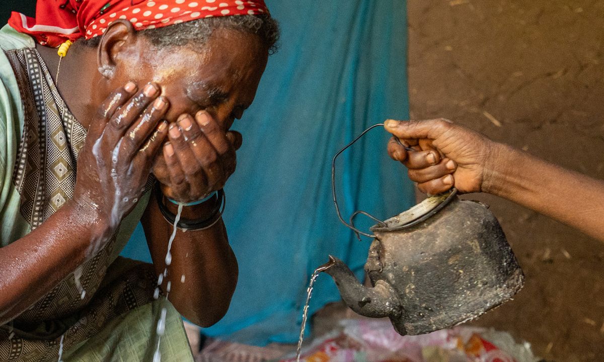 Eine Frau mit rotgepunktetem Kopftuch wäscht sich die Augen mit Wasser, das jemand aus einer kleinen Blechkaffeekanne gießt.