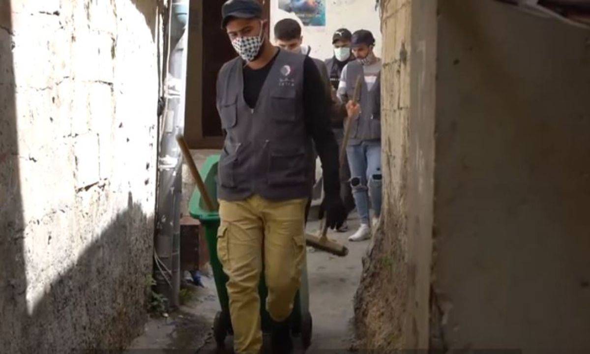 Mann mit Mund-Nasen-Schutz in einer engen Gasse zieht eine Tonne hinter sich her
