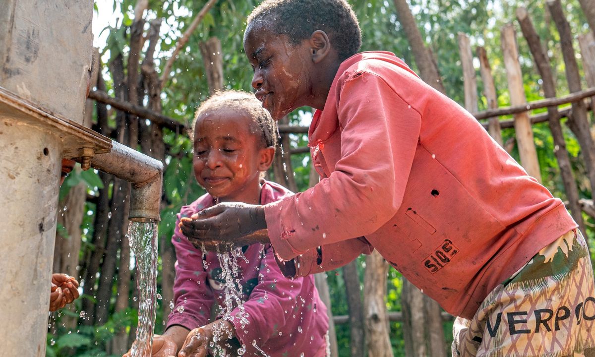 Zwei Kinder halten die Hände in den Wasserstrahl, der aus einem Brunnenrohr kommt.