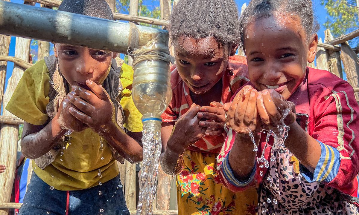 Drei Kinder neben dem Wasserstrahl eines großen Wasserhahns haben die Hände mit Wasser gefüllt.