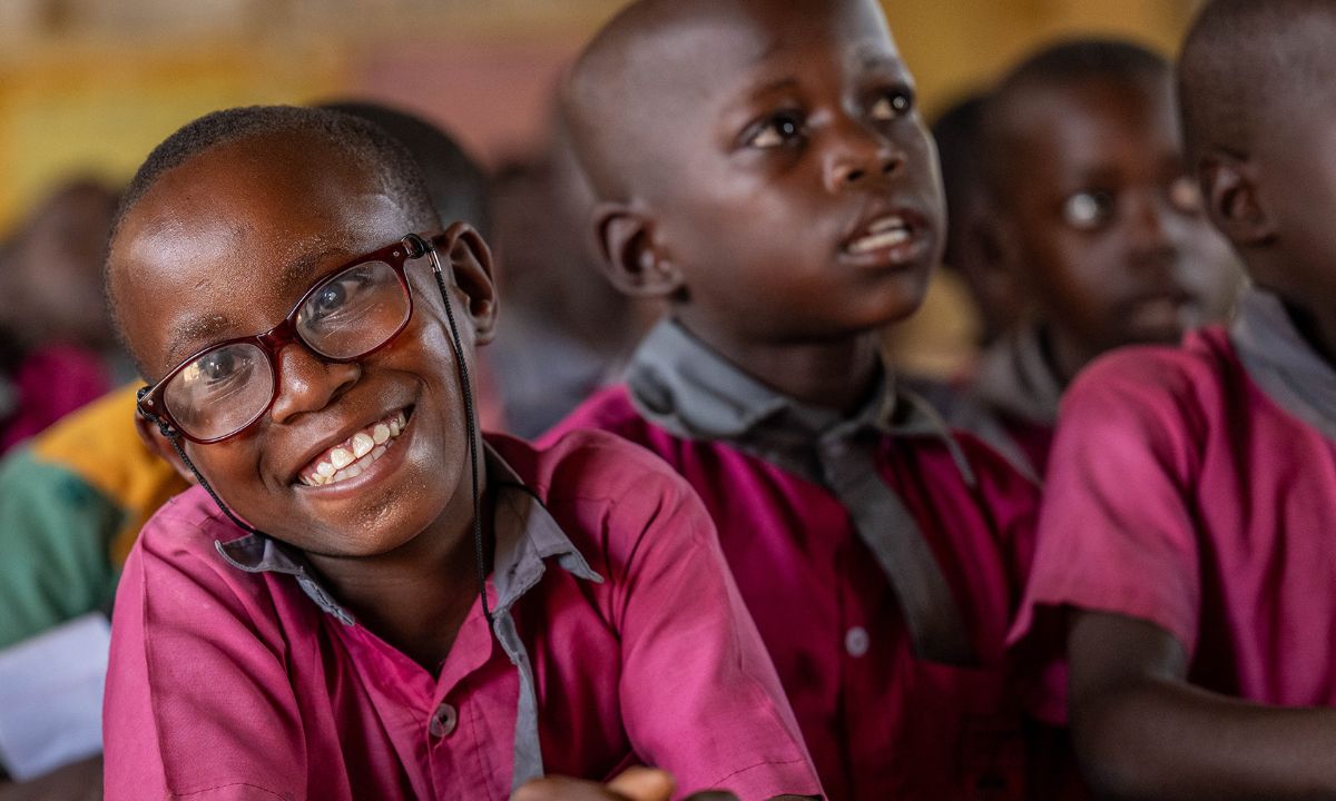 Ein lachendes Mädchen mit Brille sitzt zwischen anderen Kindern im Klassenzimmer.