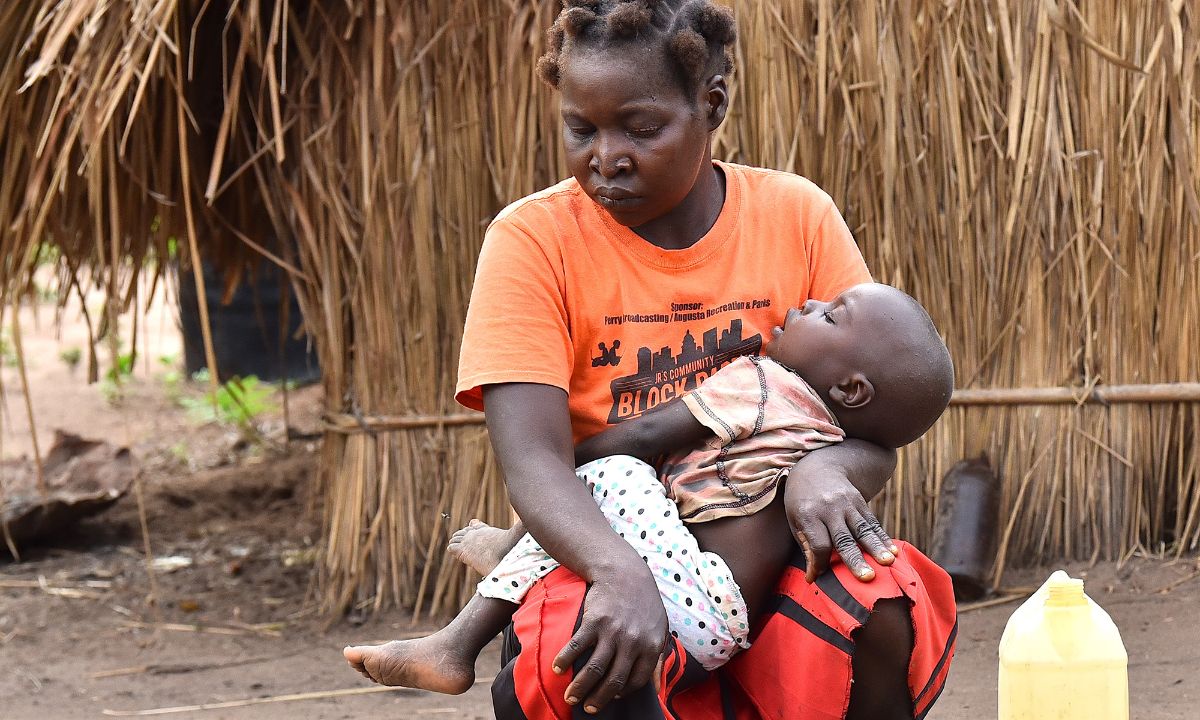 Eine Frau hält die Hand vor die Augen, sie sitzt mit einem Baby auf dem Schoß vor einer Strohhütte.